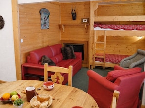 A cozy cabin interior with a red sofa, a wooden table with fruits and a mug, a bunk bed with red bedding, and a fireplace.
