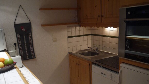 A small kitchen with a sink, stove, and dishwasher. There is a fruit bowl on the counter and a decorative item on the wall.