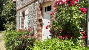 A stone building with a white door and windows, surrounded by lush greenery and pink flowers.
