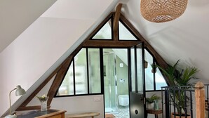 A room with a sloped ceiling, wooden beams, a desk with a lamp, a bench, and a potted plant.