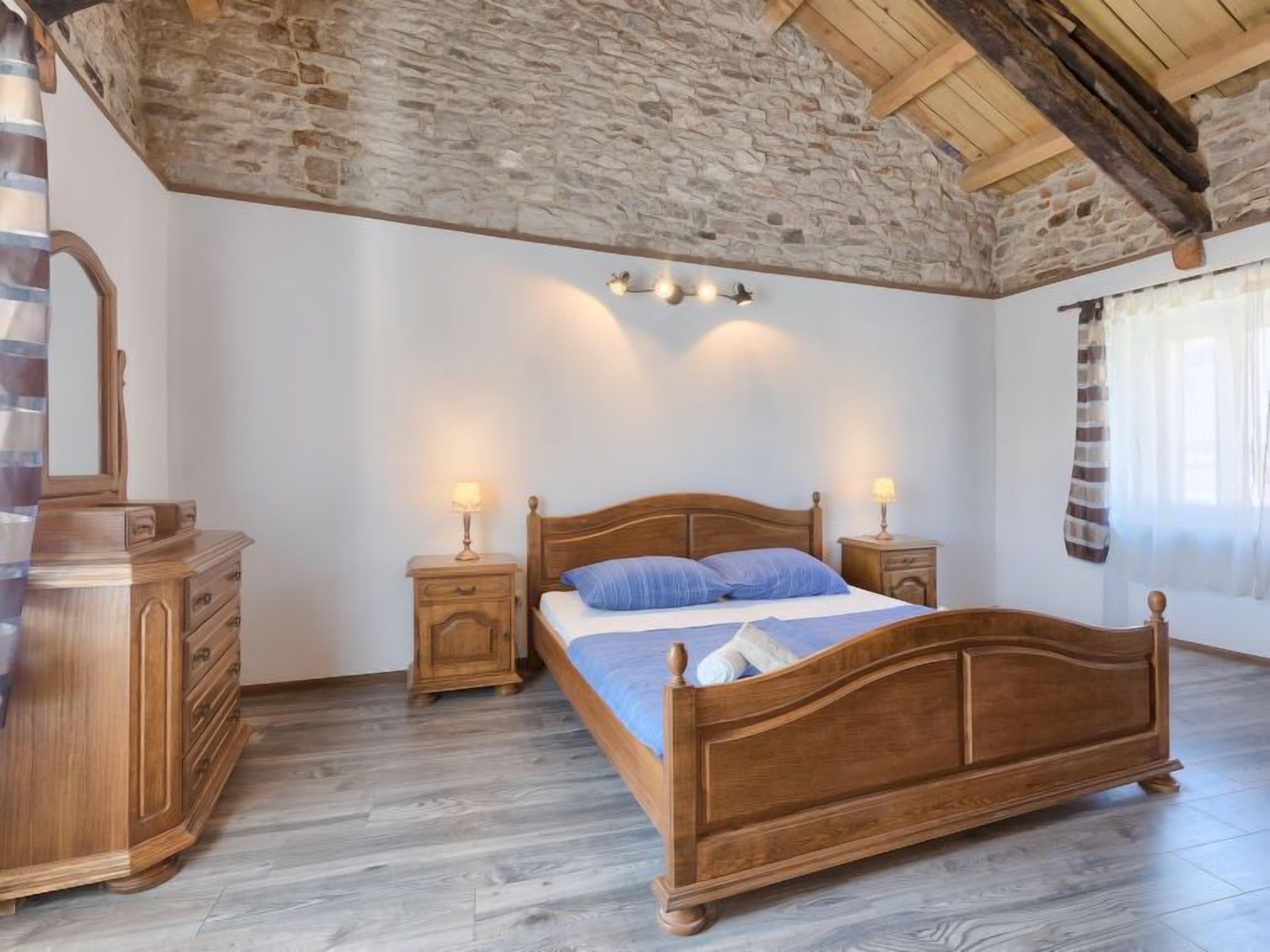 A bedroom with a wooden bed, two bedside tables with lamps, a wooden dresser, and a stone wall.