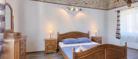 A bedroom with a wooden bed, two bedside tables with lamps, a wooden dresser, and a stone wall.