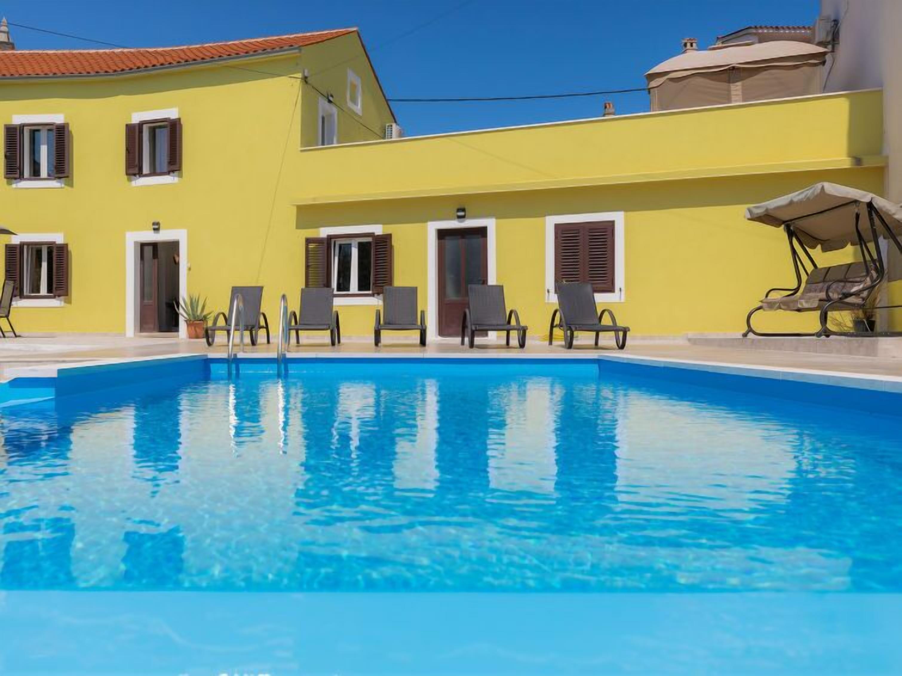 A swimming pool with lounge chairs and a yellow building with shutters.