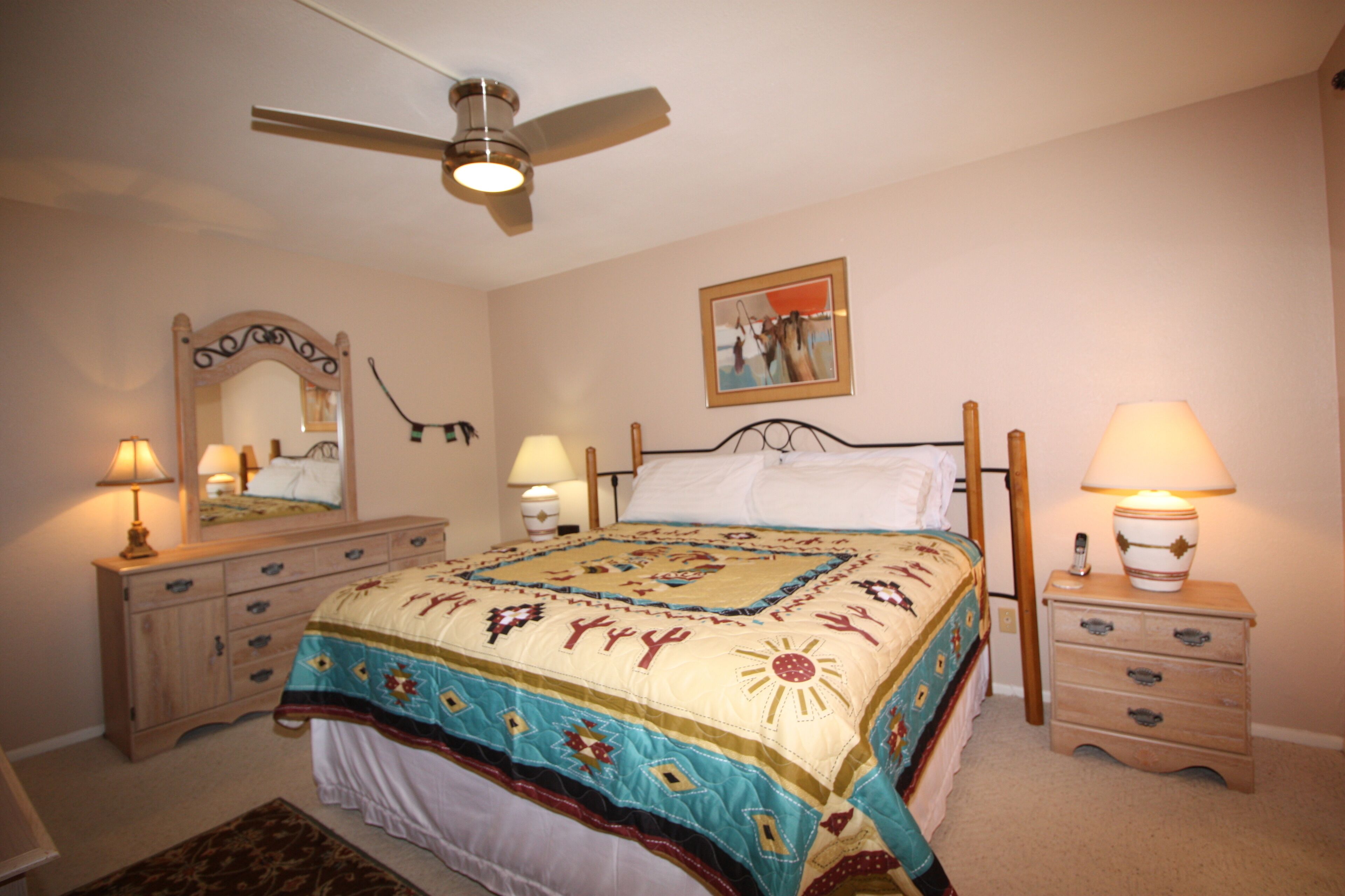 Une chambre à coucher avec un lit, deux tables de chevet avec des lampes, un ventilateur de plafond, une commode, un miroir et un tableau encadré au mur.