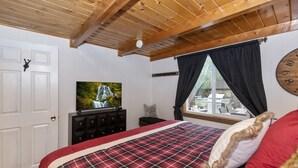 Une chambre avec un plafond en bois, un lit recouvert d’une couverture à carreaux, un téléviseur posé sur une commode et une fenêtre avec des rideaux.