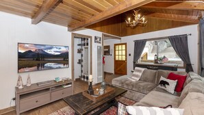 Un salon chaleureux avec un plafond en bois, un grand téléviseur fixé au mur, un canapé agrémenté de coussins décoratifs et une fenêtre avec des rideaux.