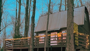 A rustic cabin with a wooden deck, surrounded by tall trees.