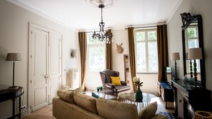 A well-lit living room with a sofa, armchair, and a table with a lamp. There is a door and a window with curtains. - Elisabeth's suite in the heart of the medieval center of Sarlat (Sarlat-la-Canéda)