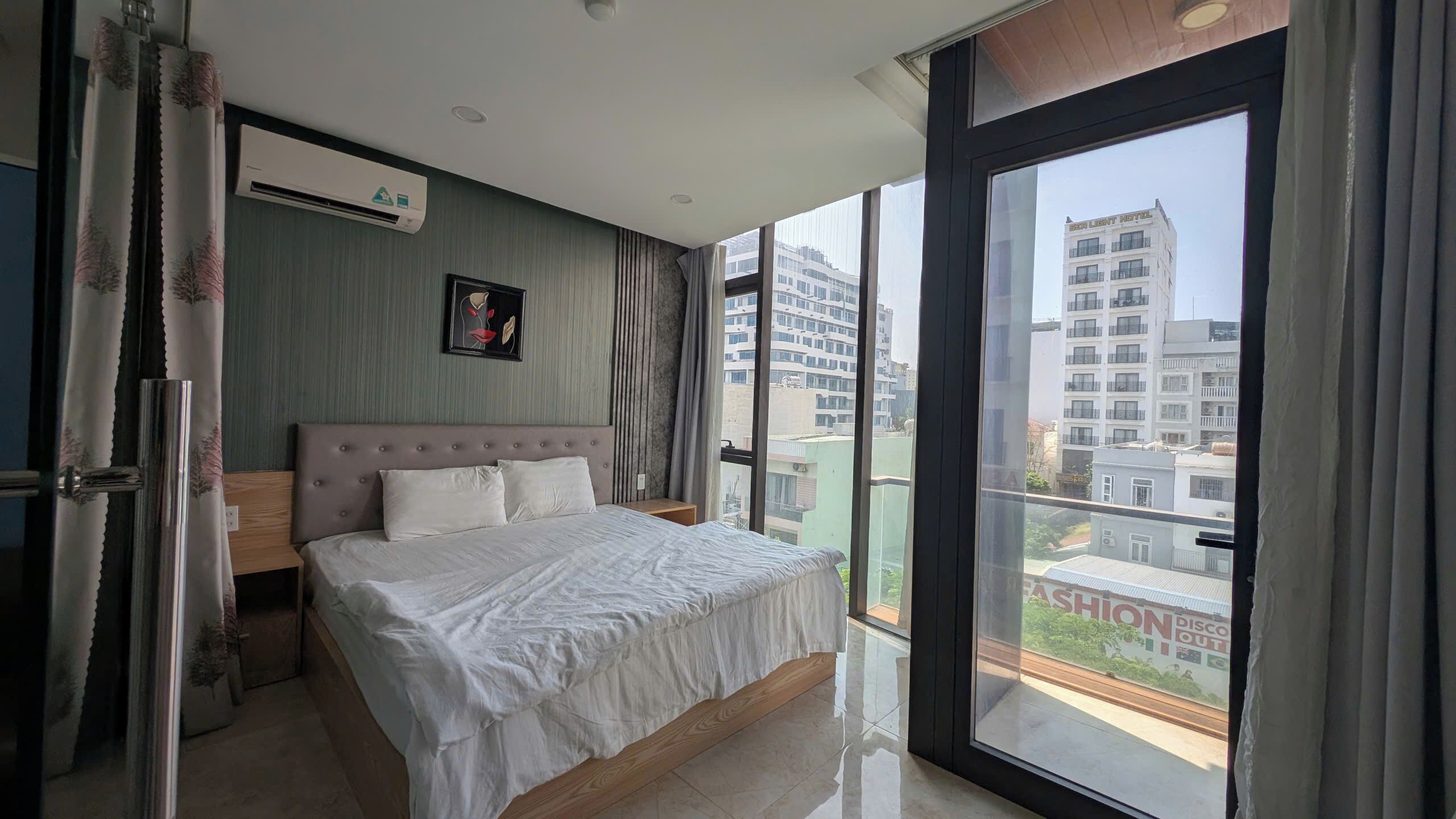 A bedroom with a bed, an air conditioning unit, a window with a view of buildings, and a curtain.