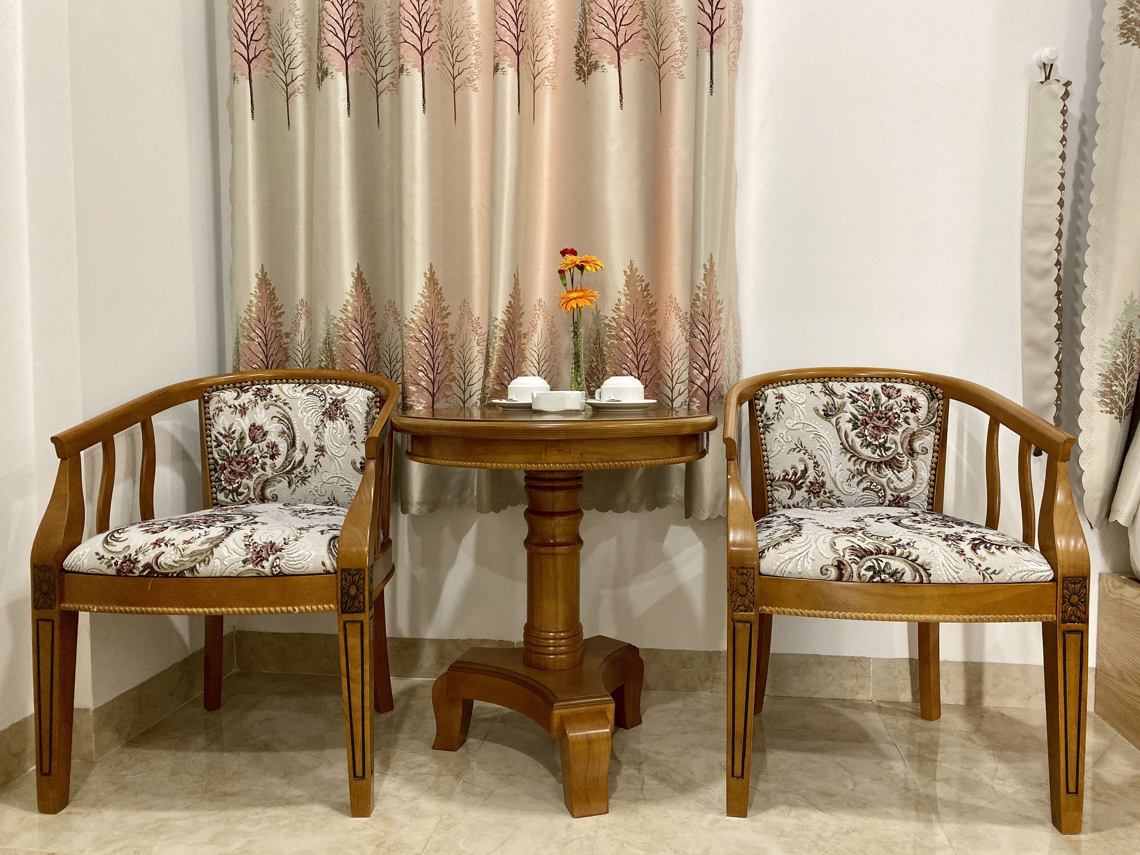 Two wooden chairs with patterned upholstery, a round wooden table, and a vase with flowers.