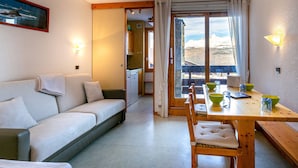 A cozy living room with a sofa, dining table, and chairs, a kitchen in the background, and a scenic mountain view through the window. - 2-room apartment, 4 people, pets allowed, Mont Blanc view (La Plagne-Tarentaise)