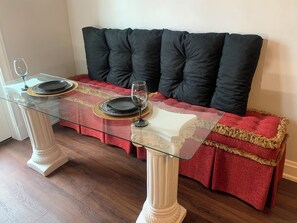 A dining table with glass top, black plates, and wine glasses, set against a red upholstered bench with black cushions.