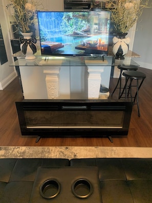 A modern living room with a glass-top coffee table, a flat-screen TV, two black barstools, and decorative vases with plants.