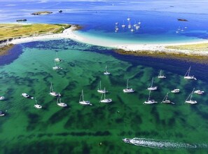 An aerial view of a coastal area with multiple sailboats anchored in clear waters, a sandy beach, and a small island in the distance.