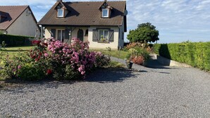 Une maison à deux étages avec une allée en gravier, entourée d’arbustes en fleurs et d’une pelouse bien entretenue.