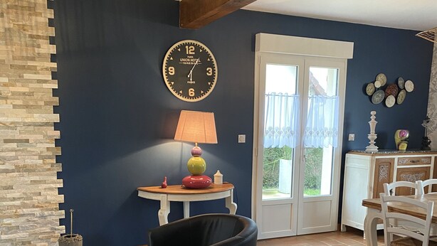 Un salon avec un mur en pierre, un fauteuil noir, une table en bois et un coin repas avec des chaises blanches.