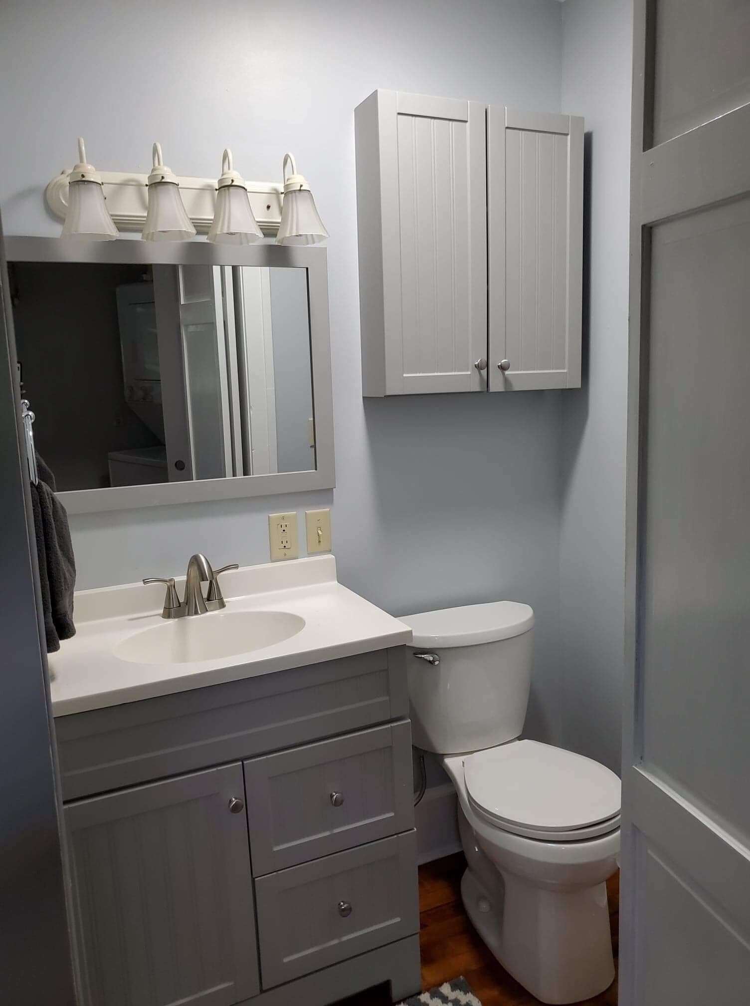 Une salle de bain avec un lavabo blanc, un miroir et des placards, des toilettes et une porte.