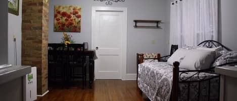 Une chambre avec un plancher en bois, un lit recouvert d’une couvre-lit à motifs, un bureau avec une chaise, un mur en briques et une porte.