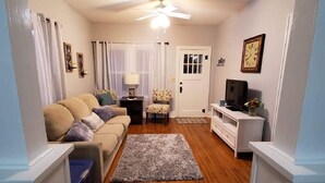 Un salon avec un canapé beige, un tapis à motifs, un ventilateur de plafond, une porte blanche et un téléviseur posé sur un meuble blanc.
