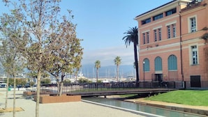 A coastal area with a building featuring arched windows and a clock tower, a paved walkway, and a marina with boats.