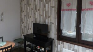 Una habitación con una televisión sobre un soporte negro, una mesita con una silla y una ventana con cortina de diseño de corazón.