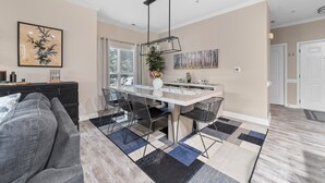 Uma sala de estar moderna com um sofá cinzento, uma área de jantar com mesa branca e cadeiras pretas, e uma cozinha ao fundo.