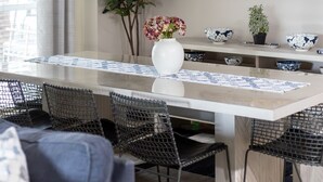 Uma área de refeições com mesa branca, cadeiras pretas, luminária pendente e uma pintura grande na parede.