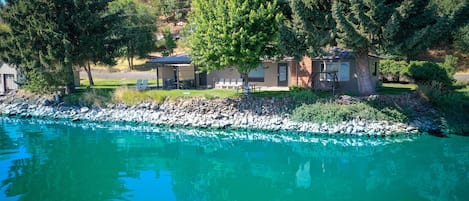 A lakeside cabin with a patio, surrounded by trees and a clear view of the water.