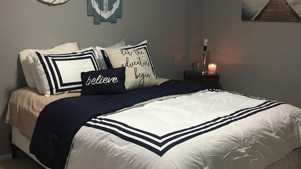 Een netjes opgemaakt bed met een marineblauw en wit dekbedovertrek, een donkerblauwe plaid en meerdere kussens, waaronder één met de tekst ‘let the adventure begin’.