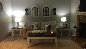 Een woonkamer met een beige bank, twee bijzettafeltjes met lampen, een salontafel met decoratieve items en een stenen openhaard.