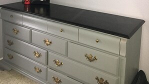 Een grijze dressoir met messing handgrepen, erboven een ingelijst kunstwerk, en een lamp met witte kap aan de linkerkant.