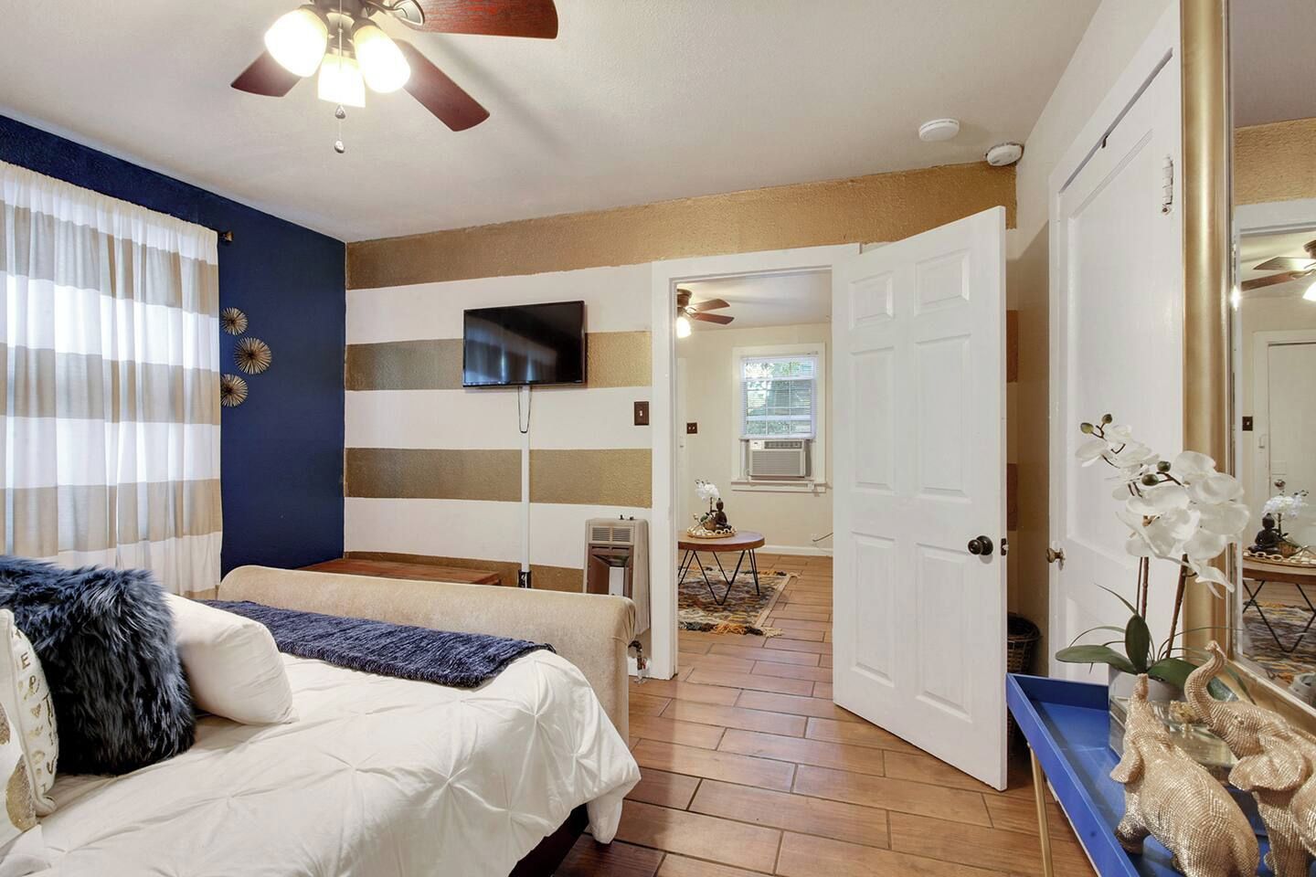 Ein Schlafzimmer mit einem Bett, einem an der Wand befestigten Fernseher, einem Deckenventilator und einer Tür zu einem weiteren Raum.