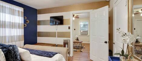 A bedroom with a bed, a TV mounted on the wall, a ceiling fan, and a door leading to another room.