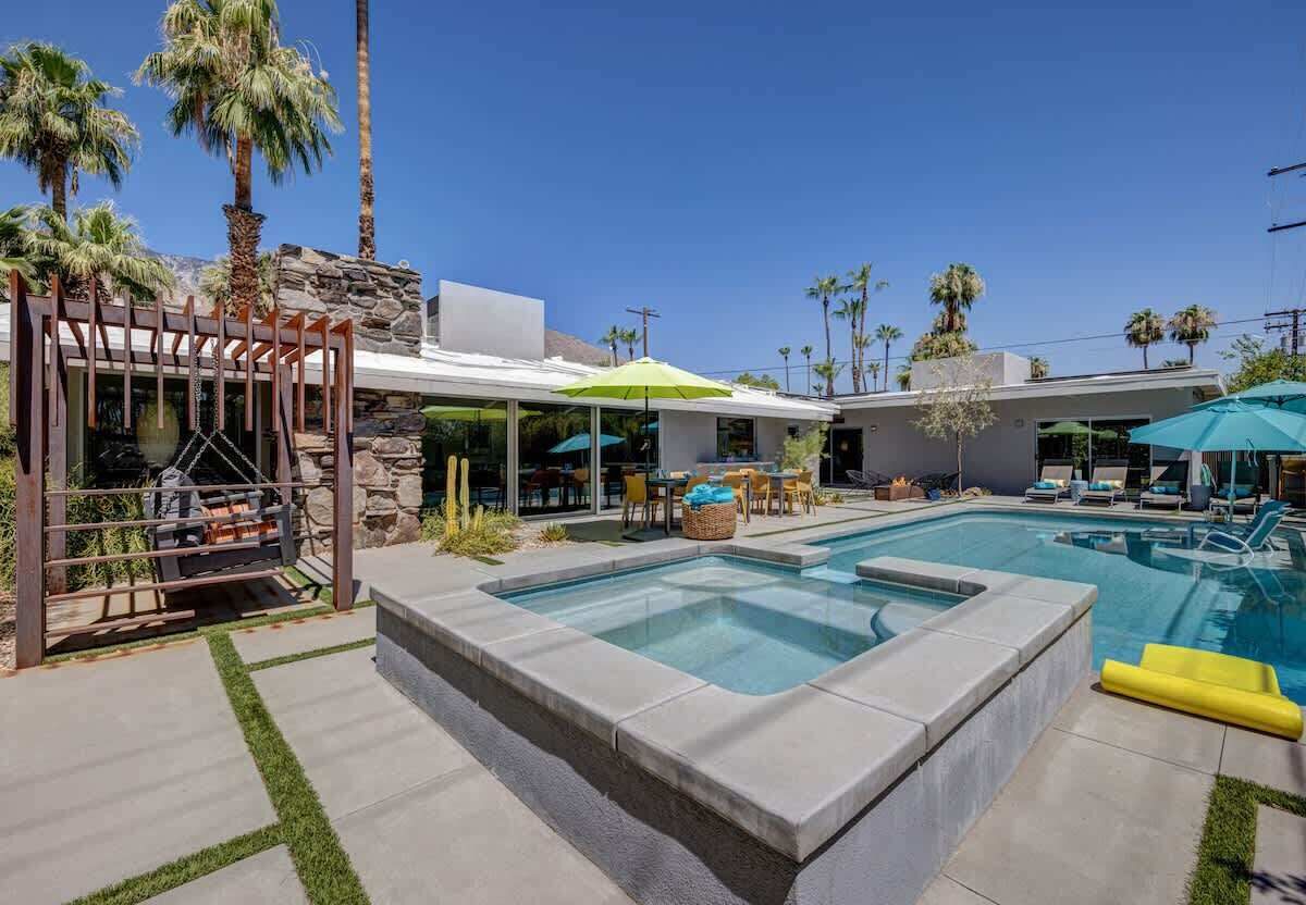 A swimming pool with a stone border, outdoor seating, palm trees, and a clear sky.