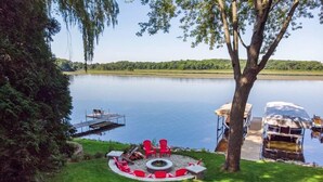 A lakeside deck with red chairs, a fire pit, and a dock extending into the water. - Blue Fish Cottage-Hot tub, Kayaks, Ice Fishing (Waterford)
