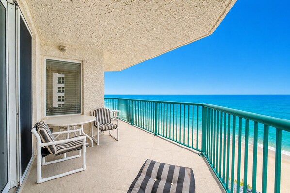 A balcony with a table and chairs, a window with blinds, and a view of the ocean.