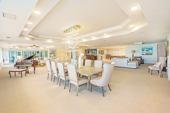 A spacious living area with a dining table, chairs, a piano, and a kitchen in the background.