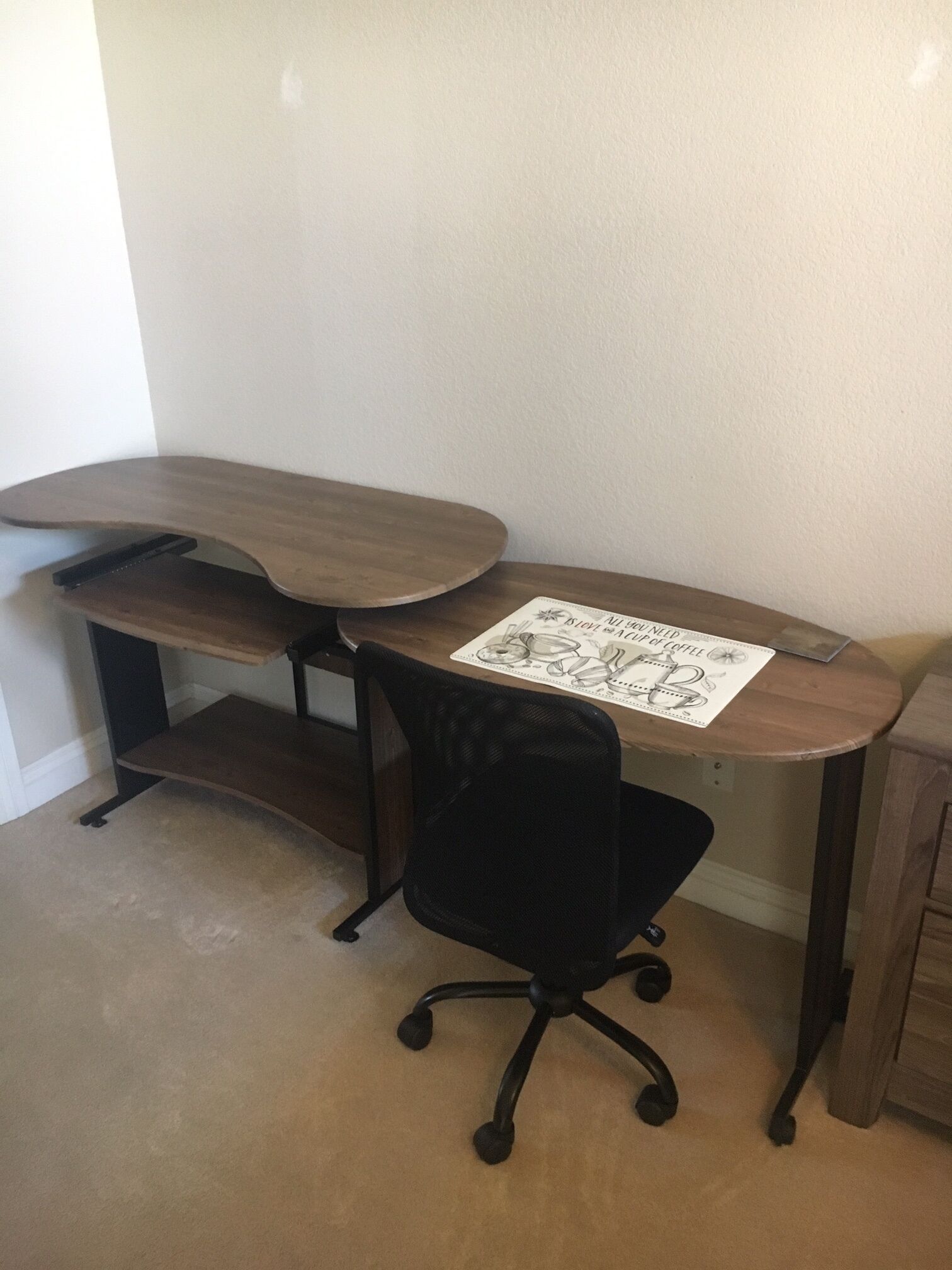 Ett skrivbord i trä med en svart kontorsstol, en vit bordsduk med ett kaffekoppsmotiv och en liten hylla.
