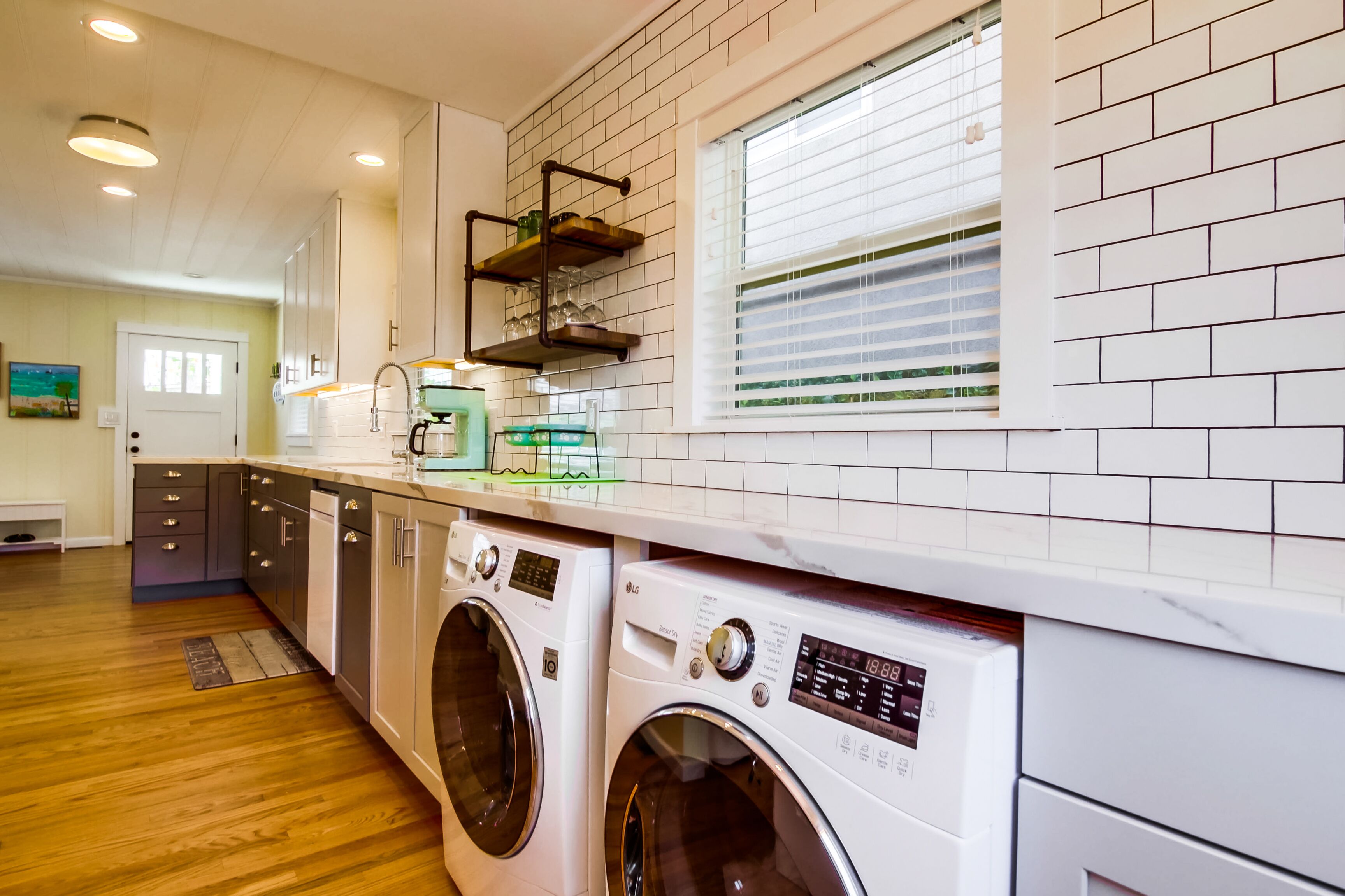 A modern laundry room with two front-loading washing machines, a countertop with a coffee maker, and a window with blinds.