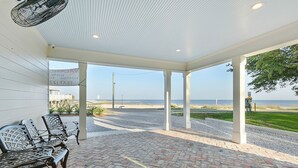 Een overdekt terras met bakstenen vloer en metalen stoelen, met uitzicht op een strand.