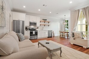 A spacious living room with a sofa, a coffee table, and a dining area with a table and chairs. The kitchen is visible with white cabinets, a refrigerator, and a microwave. - Beachfront 1 bedroom on the beach in Bay St Louis. Walking distance to Main St! (Bay St Louis)