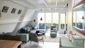 Ein gemütliches Wohnzimmer im Loft-Stil mit einer Couch, Sesseln, einem Esstisch und einem großen Fenster mit Blick auf eine Holzterrasse.