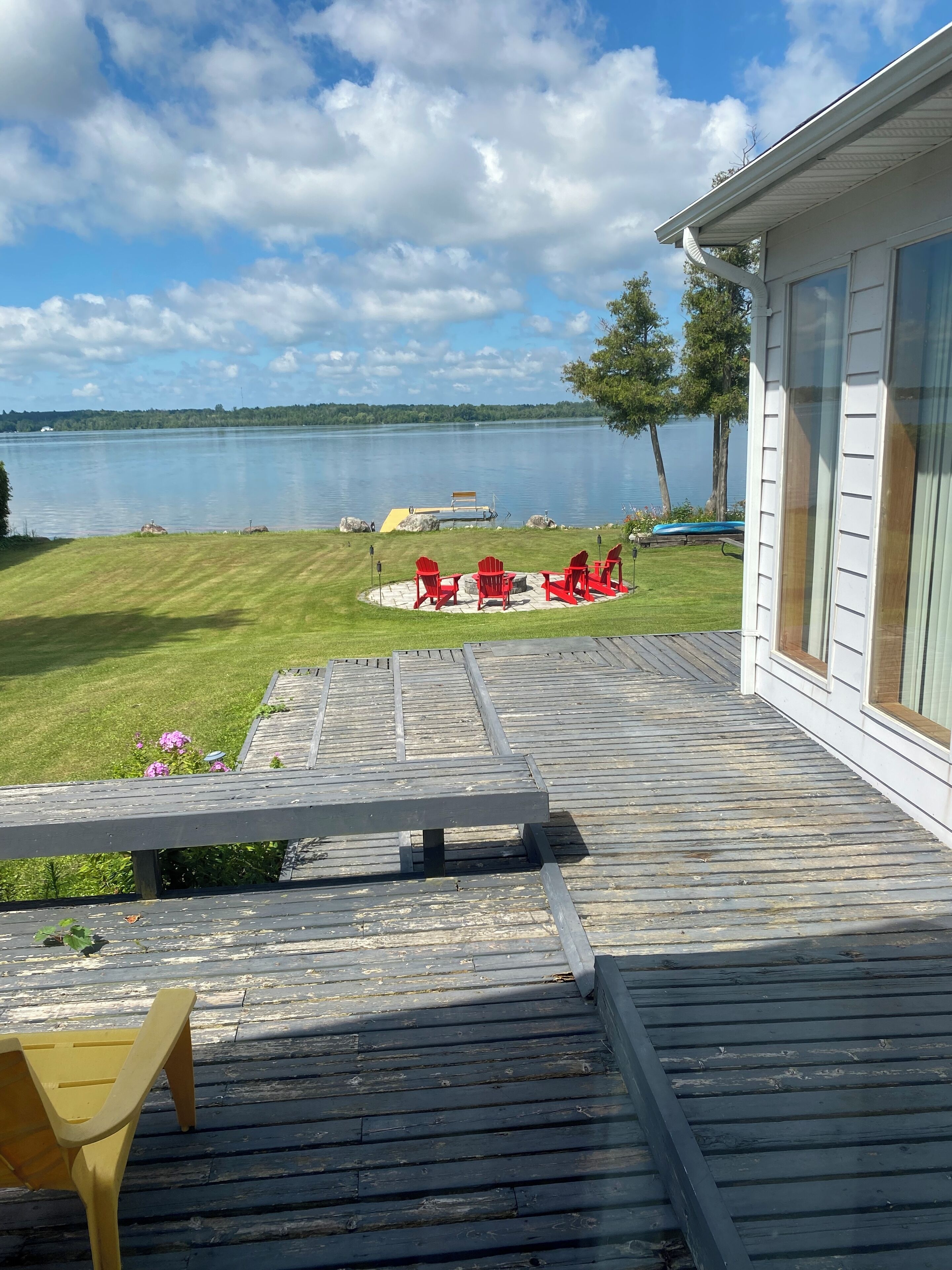 Una terraza de madera con un banco y sillas Adirondack rojas, con vista a un lago con un muelle y una zona verde.