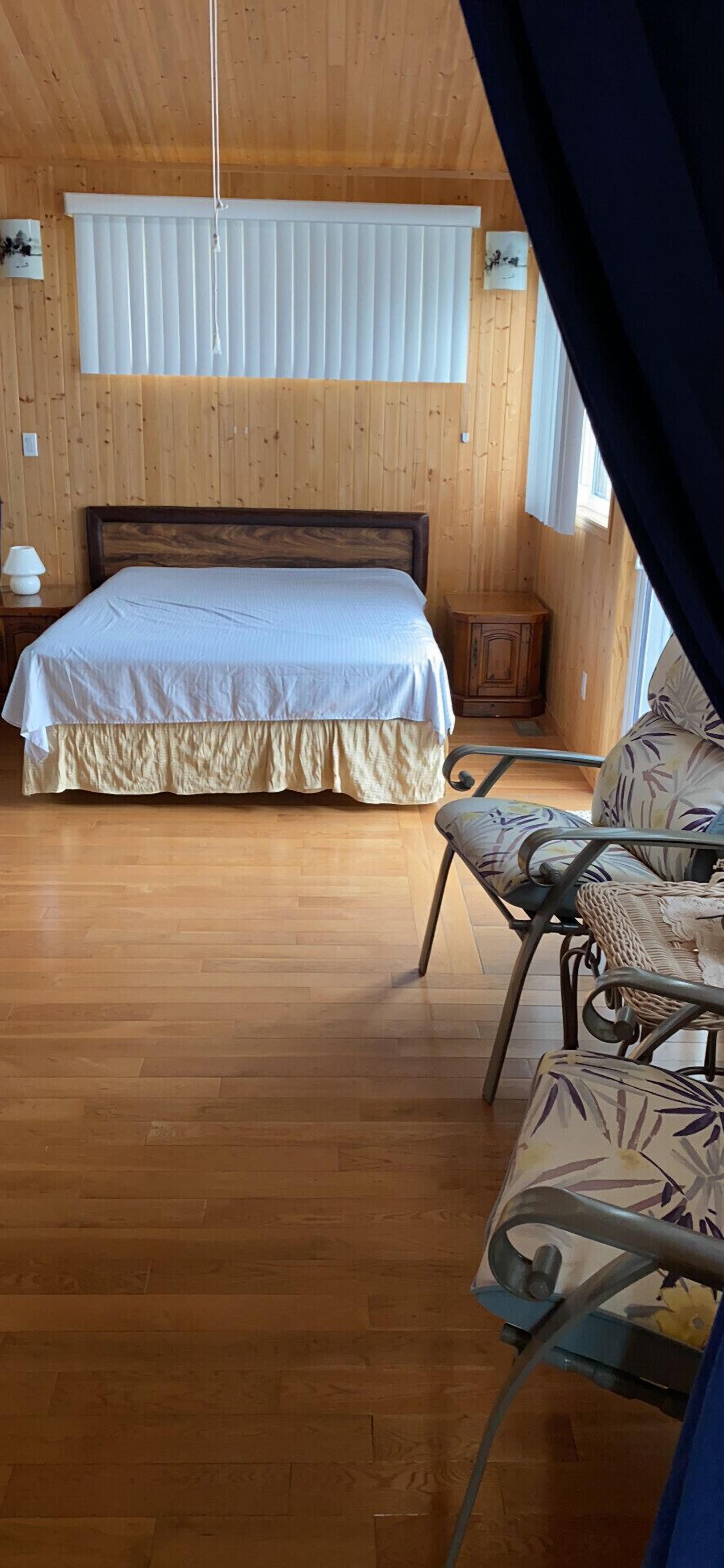 A wooden-paneled room with a bed, a small table with a lamp, and a chair with a floral pattern.