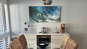 Un comedor con una mesa redonda de vidrio, dos sillas de mimbre con cojines azules, un cuadro de una ola en la pared y un armario blanco con cristalería.