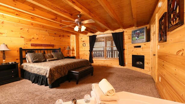 A cozy log cabin bedroom with a bed, a fireplace, a TV, and a bathroom with a bathtub. - #1  (AMAZING GATED COMMUNITY) - YOUR OWN PRIVATE (SAUNA & HOTTUB) ON BACK DECK (Sevierville)