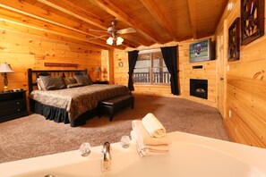A cozy log cabin bedroom with a bed, a fireplace, a TV, and a bathroom with a bathtub.