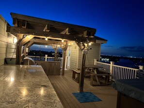 A wooden deck with a bar area, outdoor seating, and a view of the city at dusk.