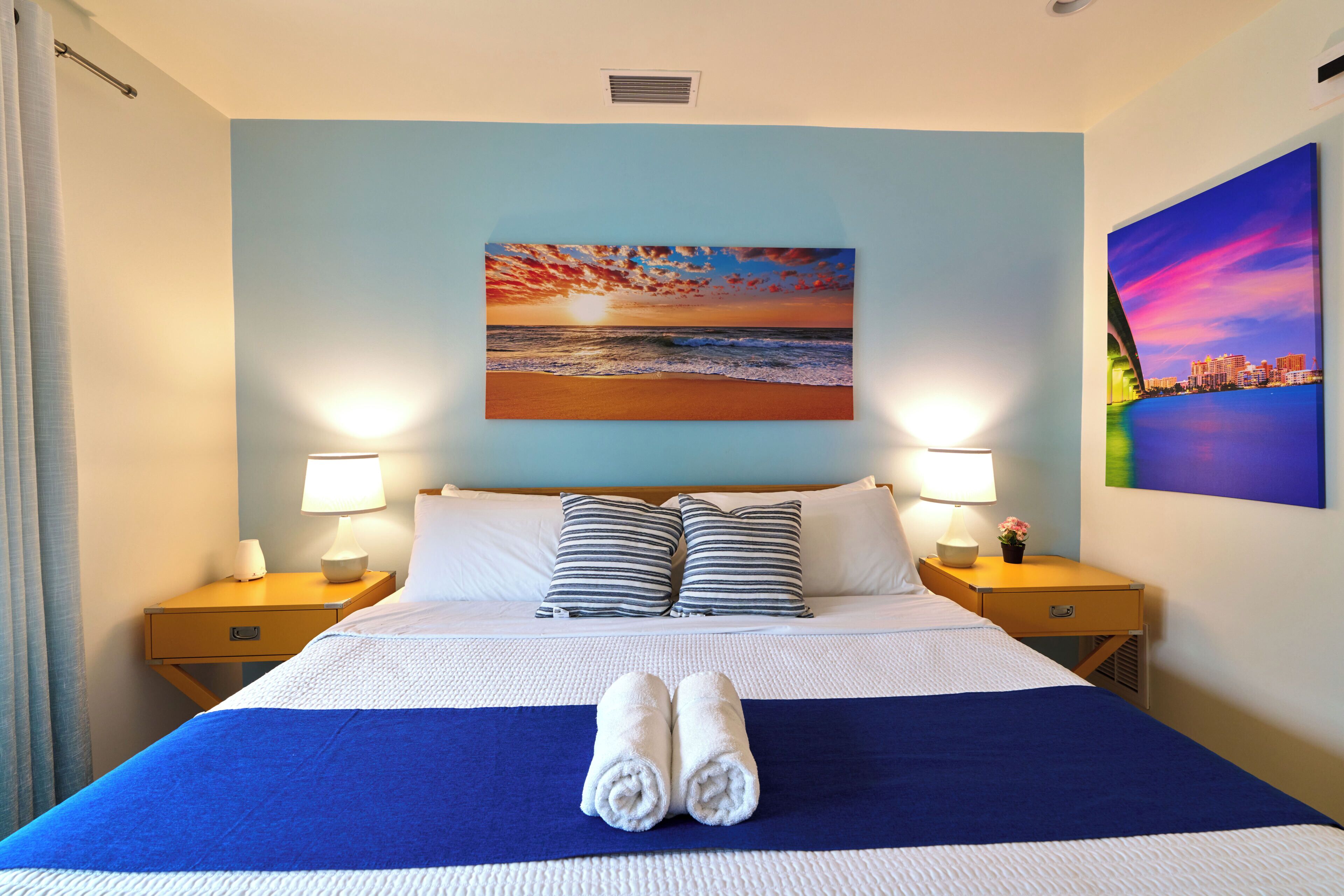 Een hotelkamer met een bed, twee bedlampen, een schilderij van een zonsondergang en een stadsgezicht.