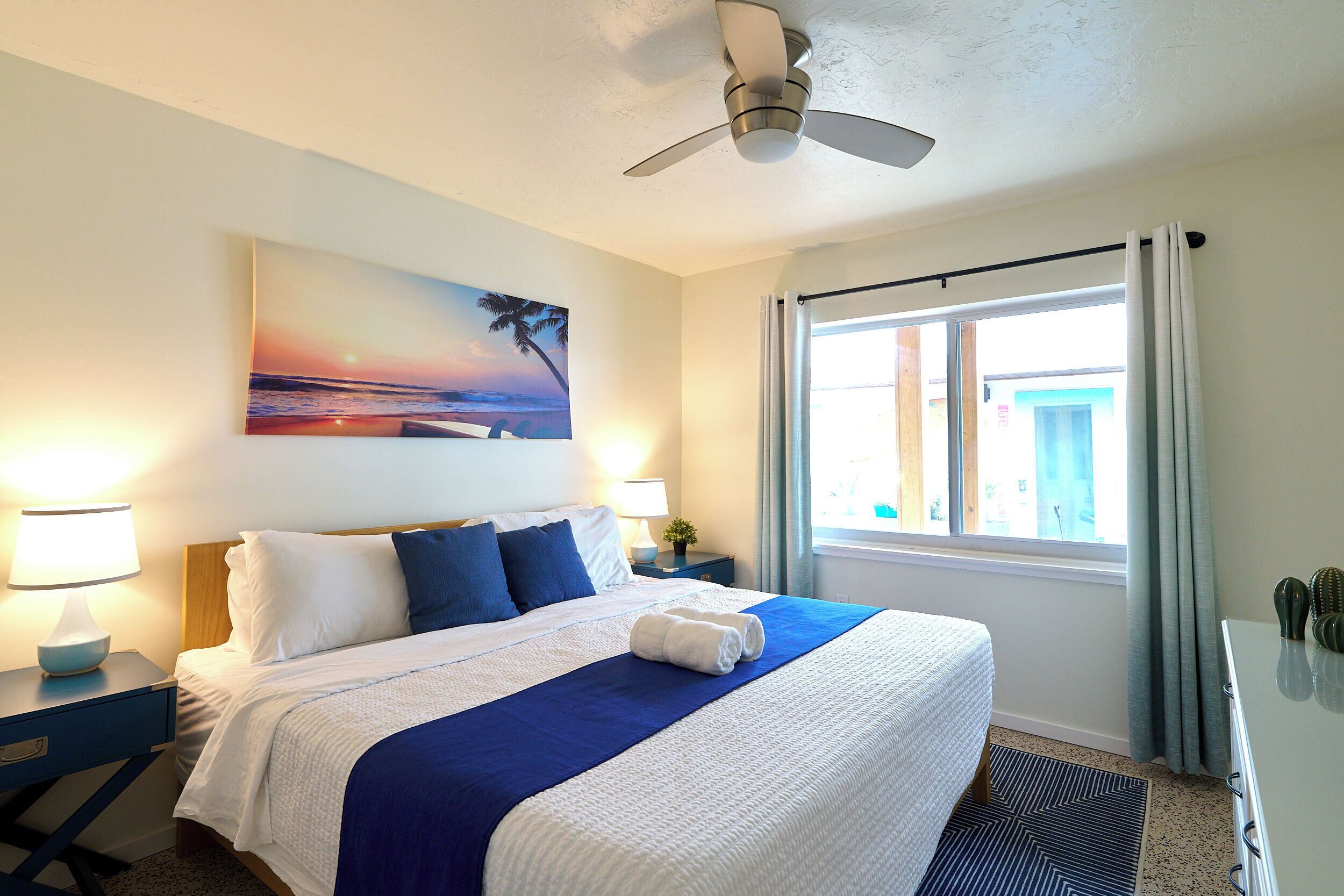 Una camera d'albergo con un letto, una ventola a soffitto, un quadro raffigurante una spiaggia, una finestra con tende e un comodino con una lampada.